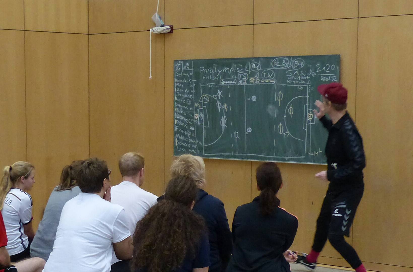 Wolf Schmidt in einer Turnhalle vor einer in gänze beschriebenen Tafel. Vor ihm sitzen auf mehreren Bänken interessierte Zuhörer.